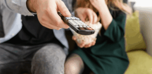 Duas pessoas sentadas em um sofá, uma segurando um controle remoto apontado para a televisão enquanto a outra segura uma tigela de pipoca.