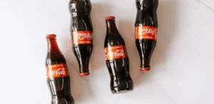 Cinco garrafas de vidro de Coca-Cola dispostas sobre uma superfície branca, algumas em pé e outras deitadas