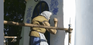 A imagem mostra uma mulher idosa pintando delicados desenhos florais e de borboletas em uma parede branca. Ela está vestindo um lenço na cabeça, um colete marrom sobre uma blusa de mangas compridas e uma saia azul com estampa de bolinhas. A mulher parece estar em um andaime improvisado, trabalhando com um pincel e um pequeno recipiente de tinta azul. Ao fundo, há uma porta azul arredondada e algumas árvores que fornecem sombra. A cena transmite um ambiente rústico e artesanal, possivelmente representando uma tradição cultural de decoração de casas.