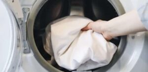 Mão colocando roupas brancas dentro de uma máquina de lavar roupa de abertura frontal.