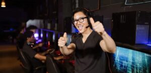 Homem sorridente usando fone de ouvido com microfone faz gesto de positivo em uma lan house, enquanto outras pessoas jogam em computadores ao fundo.