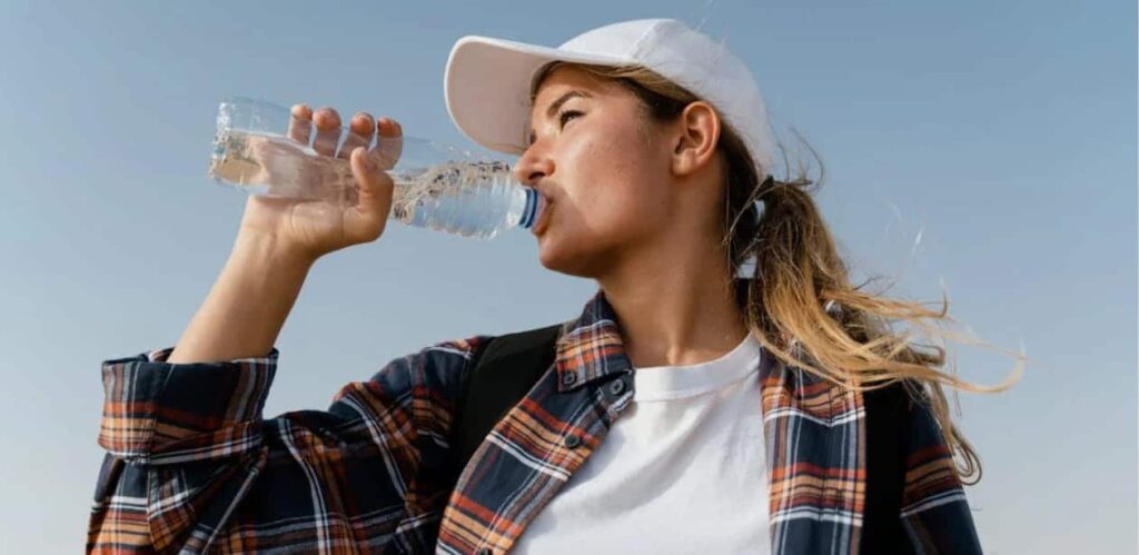 Uma jovem usando boné branco, camisa xadrez e camiseta branca bebe água de uma garrafa plástica enquanto está ao ar livre sob um céu azul. Seu cabelo loiro está preso em um rabo de cavalo, e ela carrega uma mochila preta. A imagem transmite uma sensação de calor e necessidade de hidratação.