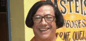 Marcos Oliveira caracterizado como Beiçola em cena da série "A Grande Família", sorrindo em frente a uma parede com letreiro de pastelaria.