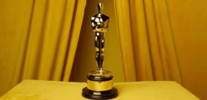 Estatueta dourada do Oscar em destaque sobre mesa amarela, com fundo de cortinas douradas.