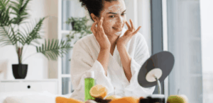 Mulher jovem aplicando máscara facial enquanto sorri para o espelho, com frutas frescas sobre a mesa