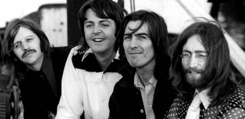 Integrantes da banda The Beatles em foto preto e branco, sorrindo e posando juntos ao ar livre