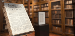 Primeiro livro de William Shakespeare, o "Primeiro Fólio", em exibição protegida em biblioteca, com estantes de livros antigos ao fundo.