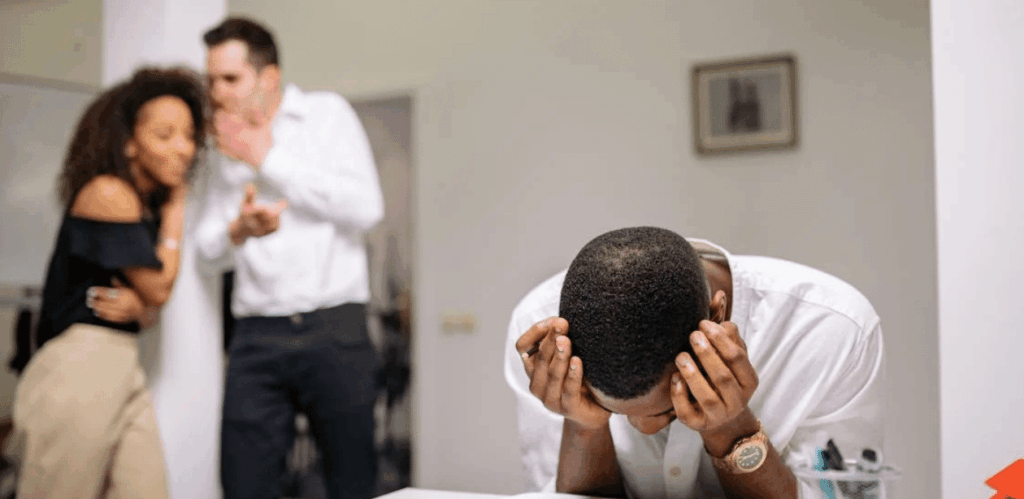 Homem com expressão de angústia em ambiente escolar, enquanto colegas ao fundo comentam em tom de deboche, representando o combate ao bullying