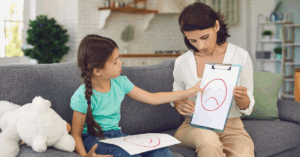 Professora mostrando desenhos de carinhas para crianças, representando emoções, em atividade de educação emocional.