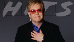 Elton John comemora sobriedade com gesto emocionado, vestindo óculos vermelhos e sorrindo diante de painel com a palavra “HITS”.