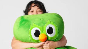 Jovem sorridente abraça uma pelúcia gigante do Duo, a icônica coruja verde do Duolingo, representando o carinho dos usuários e a febre do Duolingo no mundo.