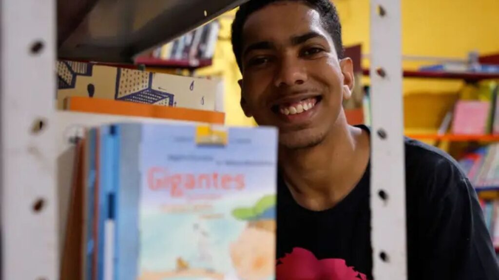 a: Jovem sorridente aparece entre prateleiras de livros em uma biblioteca pública comunitária.
