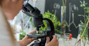 medicina integrativa - Pessoa em ambiente de laboratório observando uma amostra em microscópio. Ao fundo, há plantas em frascos de vidro e fórmulas químicas desenhadas em quadro negro, remetendo à pesquisa científica em fitoterapia e práticas integrativas.