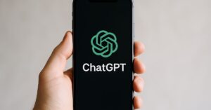 plano ChatGPT Go - Fotografia digital de uma mão humana segurando um smartphone moderno. Na tela, aparece a interface do ChatGPT, com fundo escuro e o logotipo verde característico. A iluminação suave e o fundo neutro destacam o dispositivo.