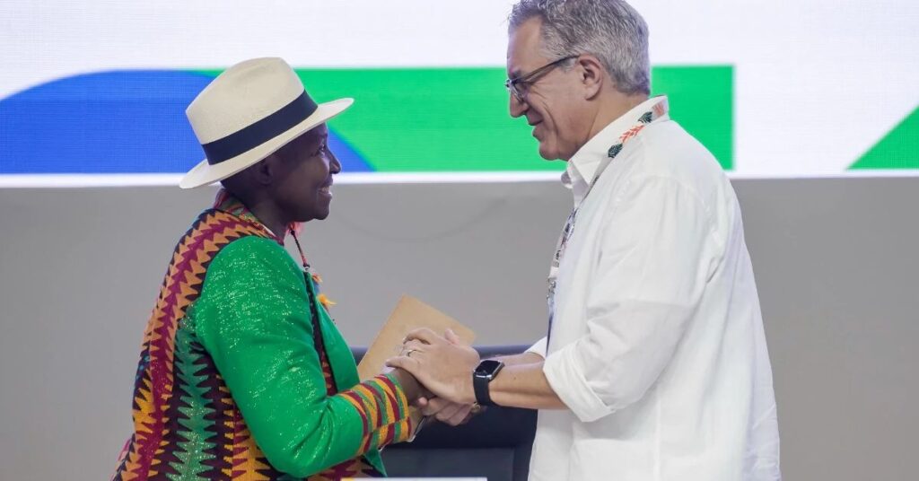 Plano de Ação em Saúde de Belém - Pessoa com chapéu claro e roupa tradicional colorida cumprimenta um representante da COP30 vestido com camisa branca, durante evento oficial em Belém.