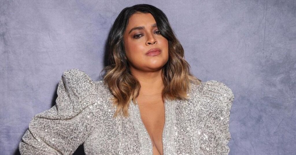 Retrato de Preta Gil usando roupa brilhante em fundo neutro.