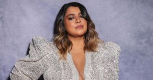 Retrato de Preta Gil usando roupa brilhante em fundo neutro.