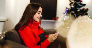 Mulher sorrindo enquanto envia frases de Feliz Natal na véspera, sentada perto da árvore decorada